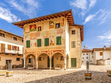 Traditionelles italienisches Haus in Spilimbergo mit bunten Fresken, grünen Fensterläden und drei Arkaden im Erdgeschoss an einem Kopfsteinpflasterplatz.