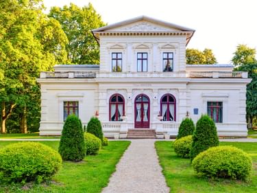 Weiße neoklassizistische Villa Genowefa mit roten Bogentüren und Fenstern, umgeben von gepflegtem Rasen und runden Sträuchern bei Heydekrug.