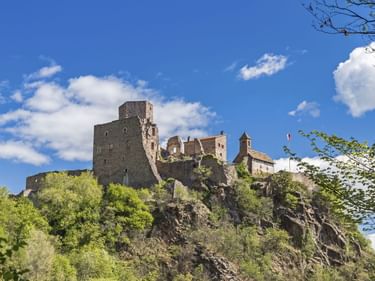 Burgruine Hocheppan mit Steinturm und Kirche auf einem felsigen Hügel, umgeben von grünen Bäumen unter blauem Himmel mit weißen Wolken.