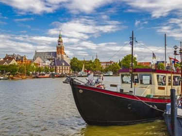 Schwarzes Boot im Hafen von Leer mit historischer Innenstadt im Hintergrund. Ein markanter Kirchturm ragt über traditionelle Gebäude unter blauem Himmel.