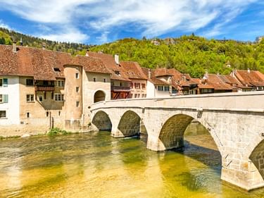 Steinbogenbrücke über Fluss in St. Ursanne mit mittelalterlichen Gebäuden, roten Ziegeldächern und bewaldeten Hügeln unter blauem Himmel.