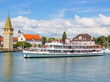 Weißes Passagierschiff Baden im Lindauer Hafen mit historischem Leuchtturm und bunten Gebäuden entlang der Uferpromenade unter blauem Himmel.