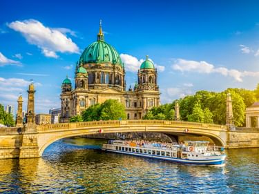 Berliner Dom mit markanten grünen Kupferkuppeln unter blauem Himmel. Ein Ausflugsschiff fährt unter einer Steinbrücke auf der Spree im Vordergrund.