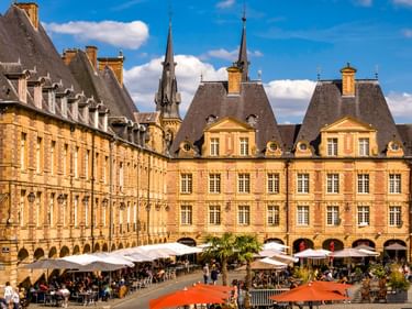 Place Ducale in Charleville-Mézières mit gelben Backsteingebäuden, steilen Schieferdächern, Arkaden und Straßencafés unter weißen Schirmen.