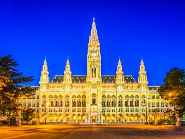 Vienna City Hall illuminated in golden light at dusk, showing its Gothic Revival architecture with central tower and spires against a deep blue sky.