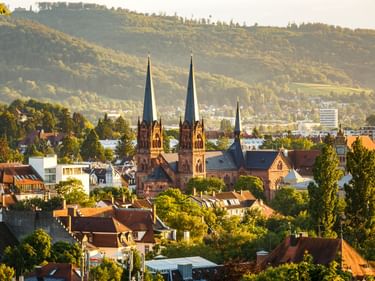 Luftaufnahme von Freiburg mit den Zwillingstürmen der Johanneskirche im Stadtbild. Rote Ziegeldächer und grüne Bäume im Vordergrund, Hügel im Hintergrund.