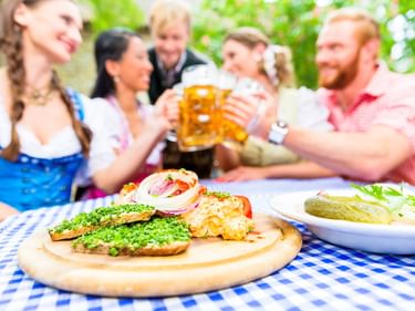 Bayerische Brotzeit mit Obatzda und Brezn auf blau-weiß karierter Tischdecke. Personen in Dirndl stoßen im Hintergrund mit Bier an.