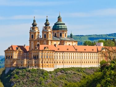 Stift Melk, ein barockes Kloster mit gelber Fassade und Zwillingstürmen mit grüner Kuppel, auf einem Felsen über dem Donautal.
