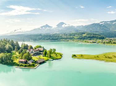 Türkisfarbener Faaker See mit kleiner Halbinsel, Häusern und grünen Wiesen. Bergkette mit Mittagskogel im Hintergrund sichtbar.