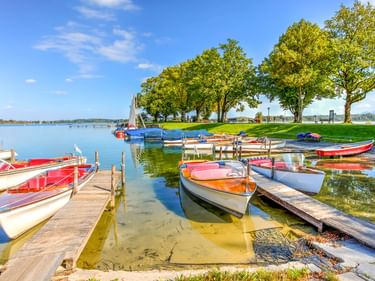Bunte Ruderboote an Holzstegen am Chiemsee in Bayern. Grüne Bäume säumen das Ufer unter blauem Himmel.