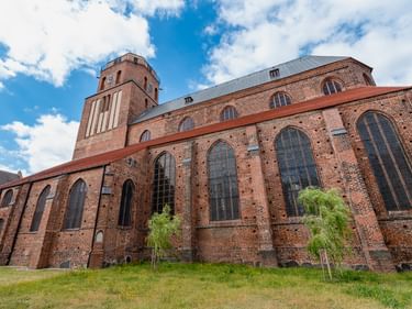 Backsteinkirche St. Petri in Wolgast mit hohem Turm und großen gotischen Fenstern, umgeben von grüner Wiese unter blauem Himmel mit weißen Wolken.