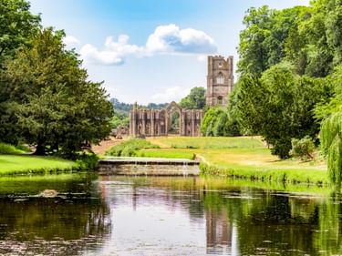 Ruinen von Fountains Abbey spiegeln sich in einem ruhigen Teich in Studley Royal, umgeben von grünen Rasenflächen und alten Bäumen.