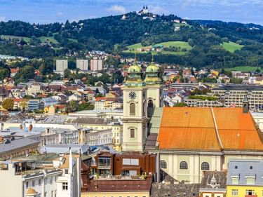 Luftaufnahme von Linz, Österreich, mit einer Kirche mit orangefarbenem Dach und grünen Kuppeln im Vordergrund und dem Pöstlingberg mit Basilika im Hintergrund.