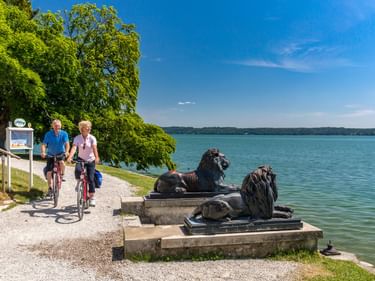 Zwei Radfahrer auf einem Uferweg am Starnberger See passieren zwei bronzene Löwenstatuen. Bäume säumen den Weg, der blaue See erstreckt sich bis zum Horizont.