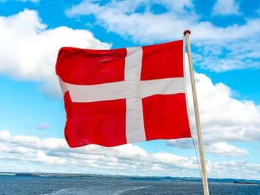 Rot-weiße dänische Flagge weht an einer Stange vor blauem Himmel mit weißen Wolken. Im Hintergrund ist eine Küstenlandschaft sichtbar.