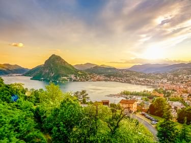 Panoramablick auf Lugano und den Luganersee mit dem Monte San Salvatore, der aus dem Wasser ragt. Grüne Hügel rahmen See und Stadt unter goldenem Abendhimmel.