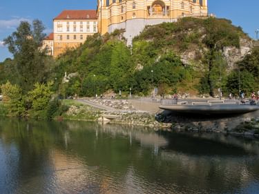 Stift Melk mit gelber Barockkirche und Klostergebäuden auf grünem Hügel über der Donau. Enten schwimmen im Vordergrund.