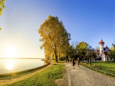 Uferweg durch Park mit Kurparkschlösschen-Turmbau in Herrsching am Ammersee. Herbstbäume rahmen den Blick auf den ruhigen See.