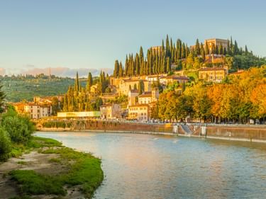 Fluss Etsch fließt durch Verona mit historischen Gebäuden am Ufer. Zypressen und buntes Herbstlaub bedecken den Hang.