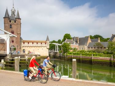 Zwei Radfahrer an einem Kanal in Zierikzee mit mittelalterlichem Turm, Zugbrücke und historischen Gebäuden, die sich im ruhigen Wasser spiegeln.