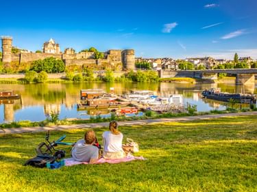 Familie beim Picknick am Flussufer mit Booten, Blick auf die mittelalterliche Festung Schloss Angers mit Türmen und Steinbrücke in Angers, Frankreich.
