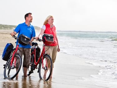 Mann und Frau mit roten Tourenrädern am Wassersaum eines Strandes in Zeeland, mit Dünen und Meer im Hintergrund.