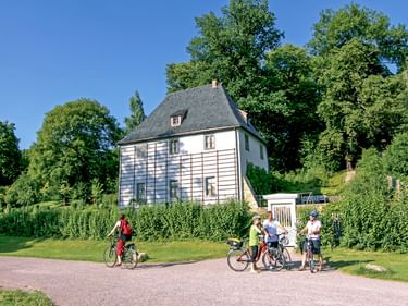 Goethes Gartenhaus, ein weißes zweistöckiges Gebäude mit dunklem Dach, umgeben von grünen Gärten. Vier Radfahrer stehen auf einem Weg vor dem Haus.