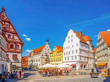 Historischer Marktplatz in Nördlingen mit bunten Fachwerkhäusern, Straßencafés und Menschen unter blauem Himmel.