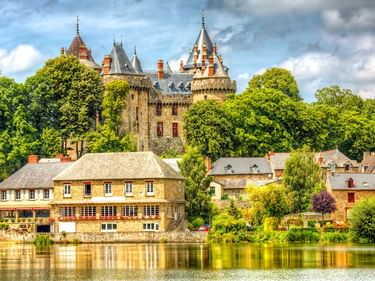 Burg Combourg mit Türmen erhebt sich über Steinhäusern an einem ruhigen See. Üppige grüne Bäume umgeben die mittelalterliche Burg unter bewölktem Himmel.