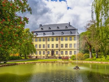 Yellow baroque palace in Gersfeld park with gray slate roof, viewed across a pond with fountain. Trees frame the building under cloudy sky.