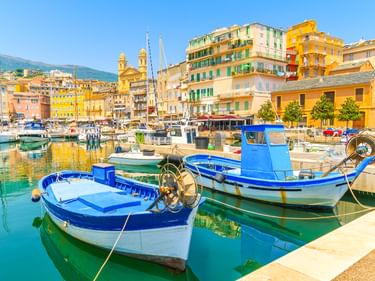 Blau-weiße Fischerboote im Vieux Port von Bastia mit bunten historischen Gebäuden und einer Kirche im Hintergrund unter blauem Himmel.