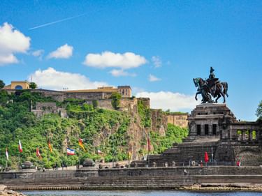 Kaiser-Wilhelm-Denkmal auf Steinplattform am Deutschen Eck in Koblenz mit bunten Flaggen, Festung Ehrenbreitstein auf grünem Hang über dem Rhein.