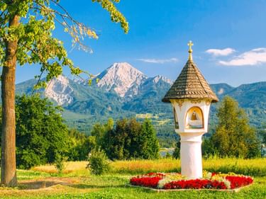 White wayside shrine with conical roof and golden cross surrounded by red flowers at Faaker See, with mountains and blue sky in background.