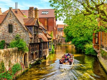 Touristenboot auf einem Kanal in Brügge, das an mittelalterlichen Backsteingebäuden mit roten Ziegeldächern vorbeifährt. Grüne Bäume säumen den Wasserweg.
