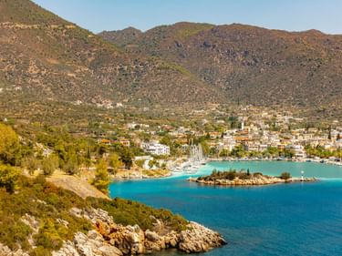 Küstenansicht von Methana, Griechenland, mit weißen Gebäuden entlang einer türkisen Bucht, umgeben von felsigen Hügeln mit spärlicher Vegetation.