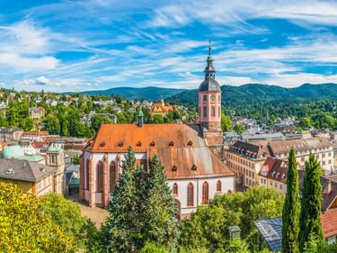 Luftaufnahme der Baden-Badener Stiftskirche mit rotem Ziegeldach und Glockenturm, umgeben von historischen Gebäuden und bewaldeten Hügeln.