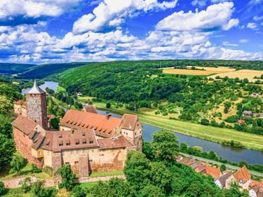 Aerial view of Rothenfels Castle with red sandstone walls and tower overlooking the Main River valley with green hills and blue sky.