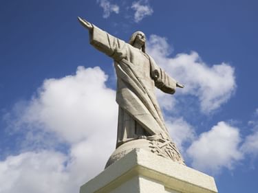 Weiße Cristo Rei Statue mit ausgestreckten Armen auf einem Sockel vor blauem Himmel mit weißen Wolken in Caniço, Madeira.