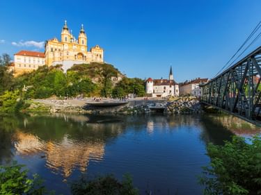 Das gelbe barocke Stift Melk auf einem Felsen über der Donau, mit einer grünen Brücke über dem Wasser und Stadtgebäuden darunter.