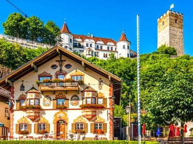 Traditionelles bemaltes bayerisches Haus mit Blumenkästen im Vordergrund, Schloss Neubeuern mit weißen Mauern und roten Dächern sowie Steinturm auf Hügel.