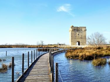 Holzsteg zur Tour Carbonnière, einem mittelalterlichen Steinturm, umgeben von Sumpfland und Wasser unter blauem Himmel.