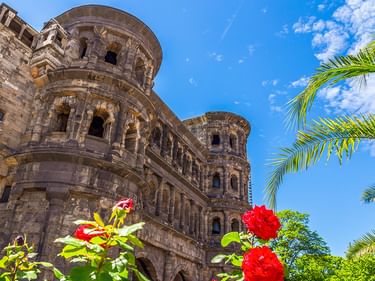 Das antike römische Stadttor Porta Nigra in Trier mit seinen charakteristischen Sandsteinbögen und Türmen. Rote Rosen und Palmwedel rahmen die Ansicht.