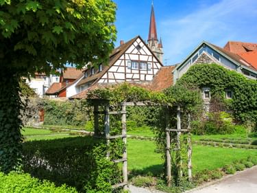 Grüner Park in Radolfzell mit Holzpergola, gepflegten Rasenflächen, Fachwerkhäusern und rotem Kirchturm unter blauem Himmel.