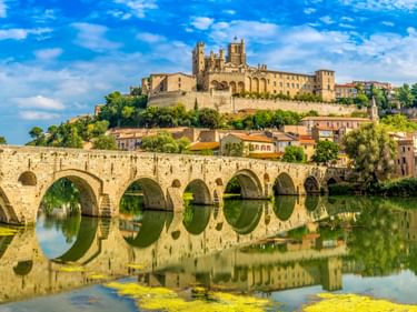 Historische Steinbrücke mit mehreren Bögen über einem ruhigen Fluss in Béziers, die sich im Wasser spiegelt. Kathedrale und Altstadt auf Hügel.