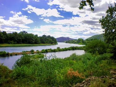 The Rhône River near Meximieux with lush green vegetation along its banks, calm blue water, and hills in the distance under a cloudy sky.