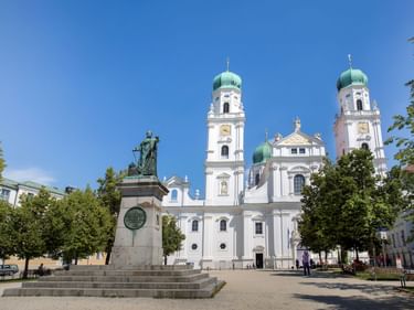 Weißer barocker Stephansdom mit zwei grünen Zwiebeltürmen und einem Denkmal im Vordergrund auf einem sonnigen Platz in Passau.