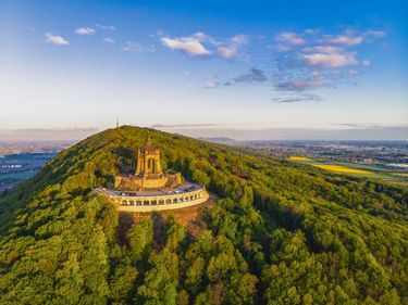 Luftaufnahme des Kaiser-Wilhelm-Denkmals auf bewaldetem Hügel in Porta Westfalica mit Flusstal und Ebenen unter blauem Himmel.