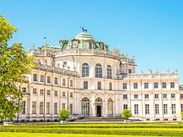 Weißes Barockschloss Stupinigi mit verzierten Balustraden und grüner Kuppel, davor gepflegter Rasen und Hecken unter blauem Himmel.