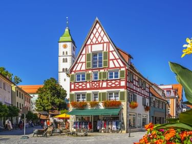 Historischer Saumarkt in Wangen im Allgäu mit buntem Fachwerkhaus, Kirchturm mit grünem Dach und Blumenkästen unter blauem Himmel.