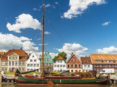 Traditionelles Segelschiff im Hafen von Glückstadt mit bunten historischen Häusern entlang der Uferpromenade unter blauem Himmel mit weißen Wolken.